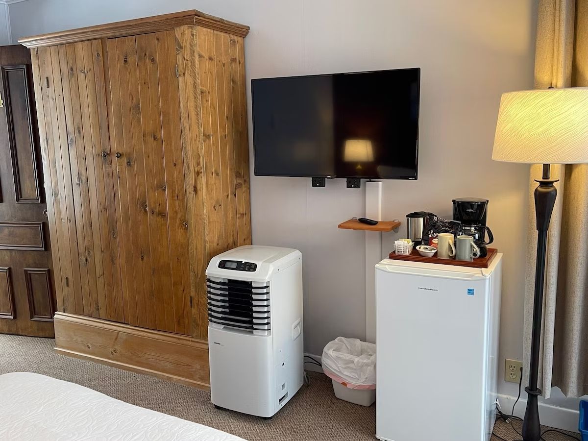 Kleine kamer met een houten kast, aan de muur gemonteerde tv, draagbare airco, minikoelkast en staande lamp.