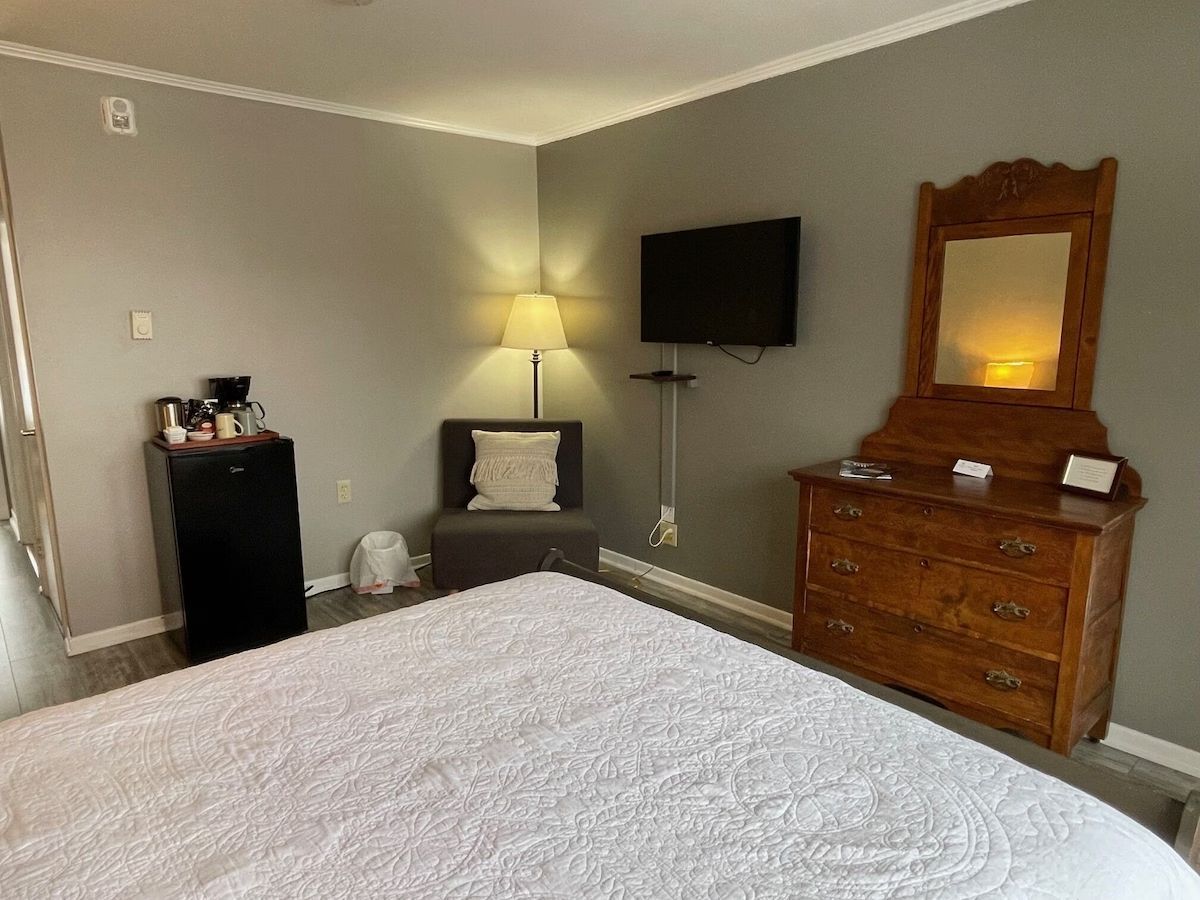 Slaapkamer met een bed, commode, lamp, aan de muur gemonteerde tv en stoel in neutrale tinten.
