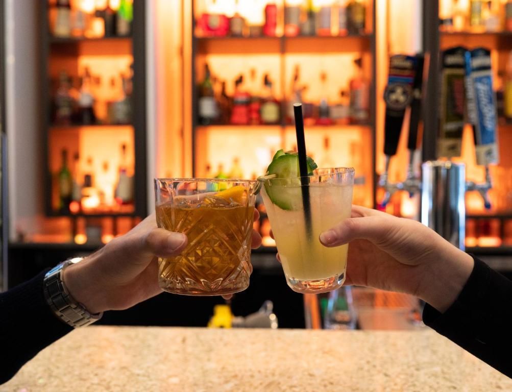 Twee handen klinken cocktails tegen elkaar aan een sfeervol verlichte bar met planken vol sterke drank op de achtergrond.