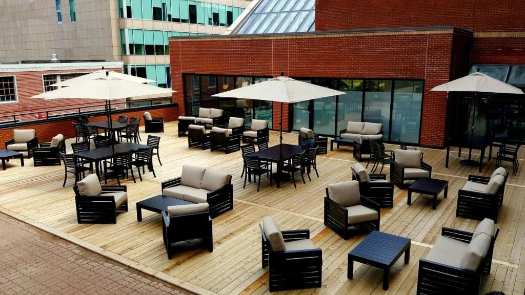 Dakterras van The Prince George Hotel, Halifax in de buitenlucht met zwarte rieten zitmeubels, tafels en witte parasols, naast moderne bakstenen gebouwen.