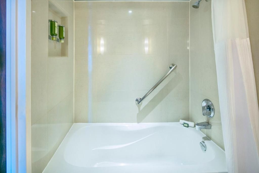 Witte badkuip en douche in een kleine, beige betegelde badkamer van The Prince George Hotel, Halifax met een aan de muur bevestigde handgreep.