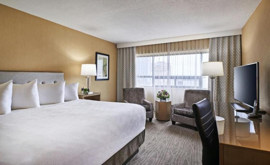 Hotelkamer van The Prince George Hotel, Halifax met kingsize bed, bureau, fauteuil, tv en groot raam met gordijnen.