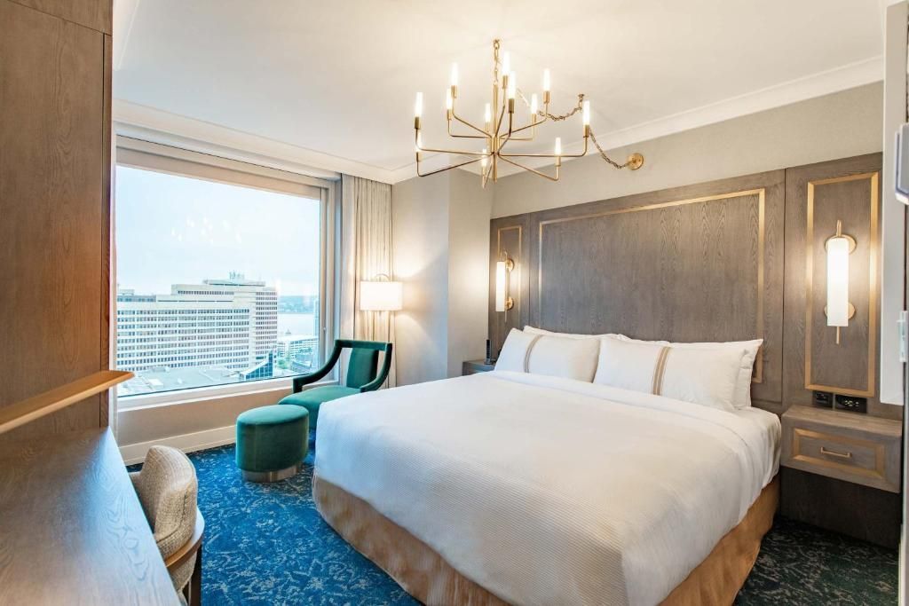 Moderne hotelkamer met een groot bed, kroonluchter, turquoise stoel en uitzicht over de stad door kamerhoge ramen.
