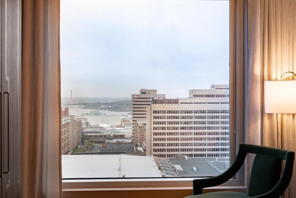 Uitzicht vanuit het hotelkamerraam op de skyline van de stad en het water bij zonsondergang, met een stoel bij het raam.