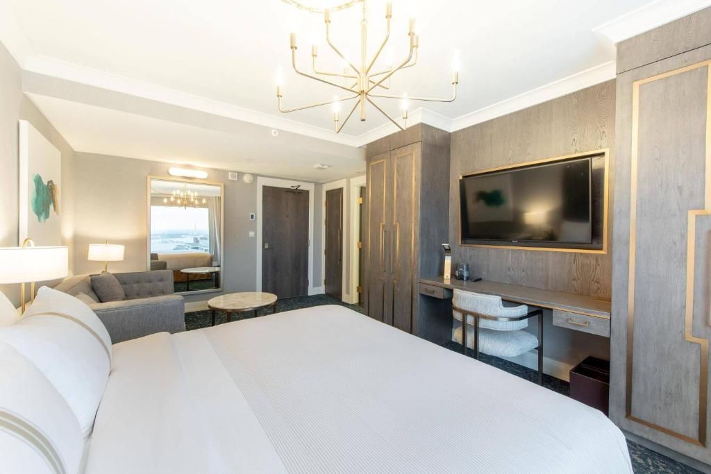 Lichte hotelkamer met kingsize bed, bank, tv, kledingkast met spiegel en kroonluchter.