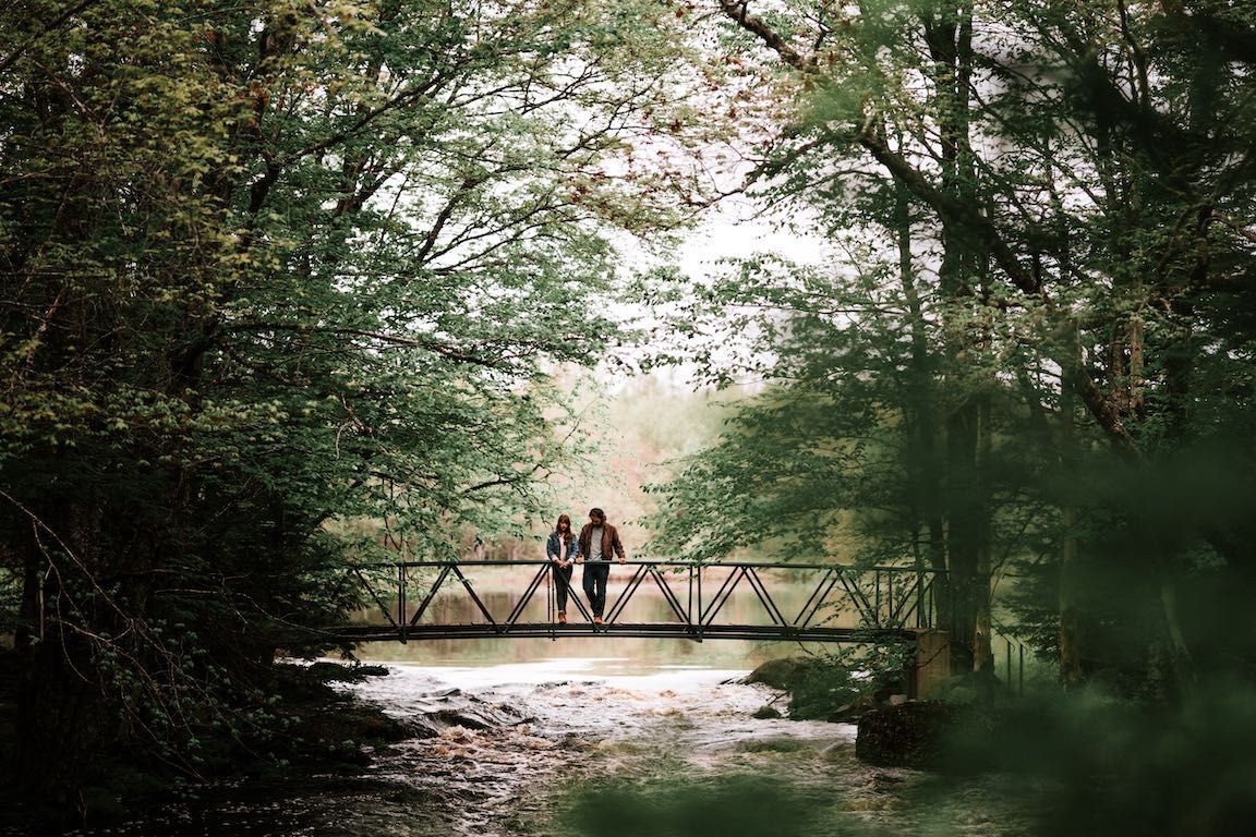 Twee mensen staan op een klein houten bruggetje in een weelderig groen bos, boven een ondiepe beek.