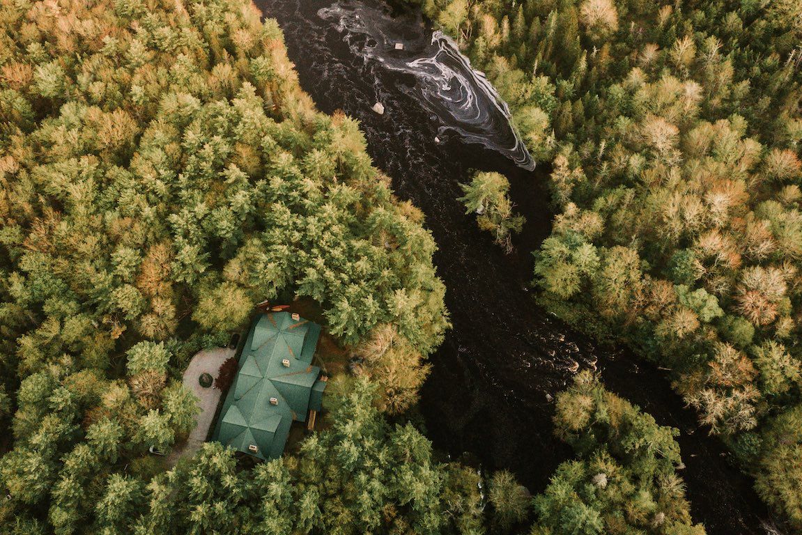 Luchtfoto van een hut met een groen dak aan een donkere rivier in een dicht herfstbos.