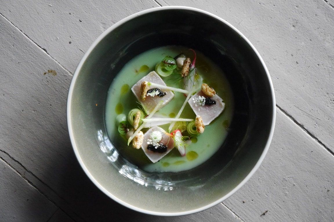 Bovenaanzicht van een zwarte kom met groene soep en gegarneerde dumplings op een grijze ondergrond.