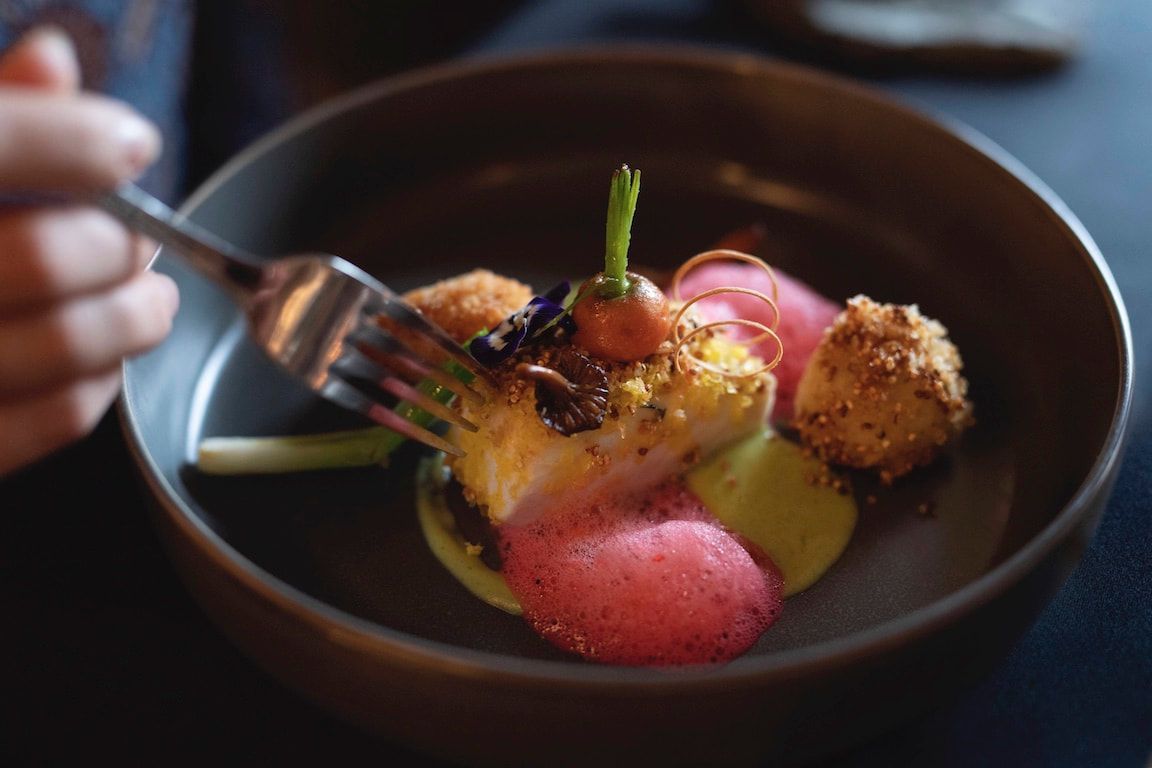 Een vork boven een kleurrijk opgemaakt dessert in een donkere kom, met roze en gele bolletjes en garnering.