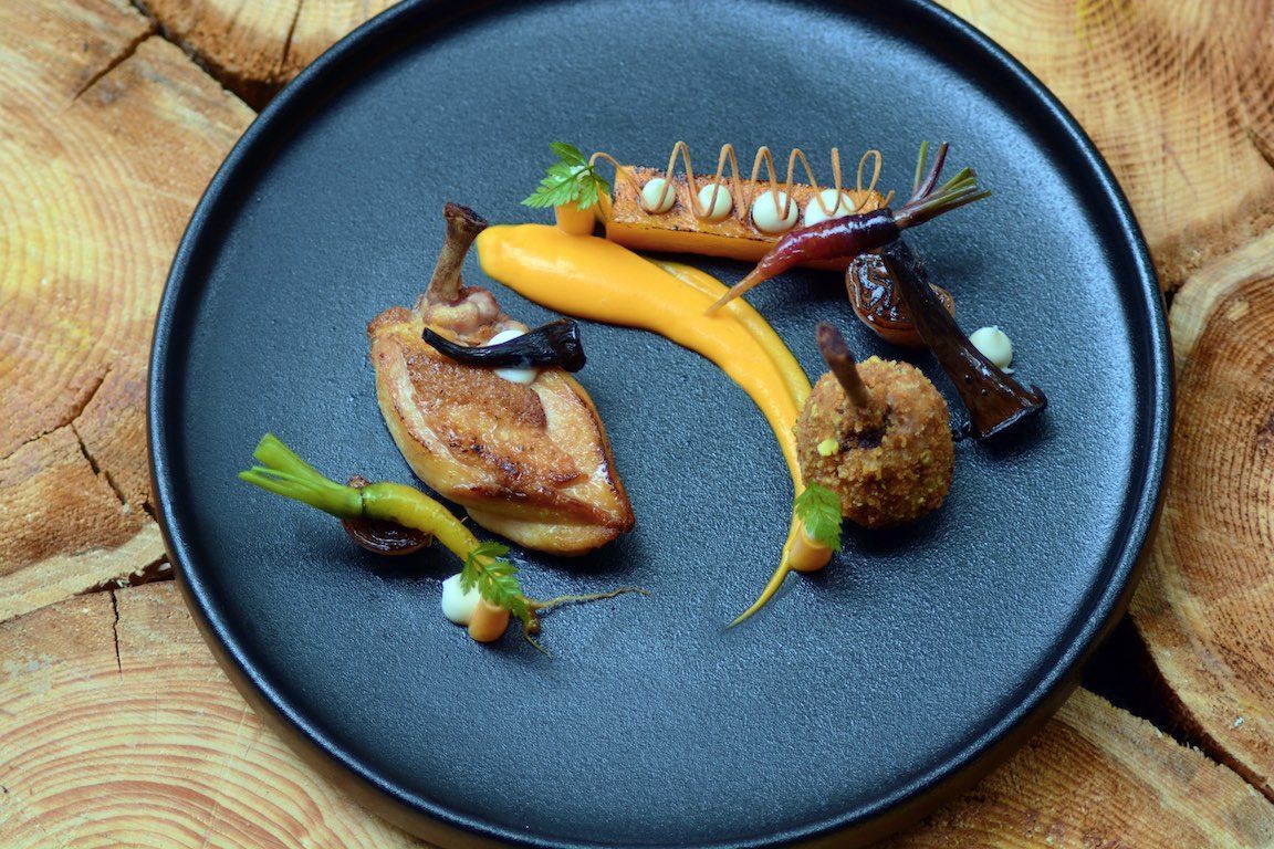 Een gastronomisch gerecht, geserveerd op een zwart bord, met saus, groenten en garnering op een houten tafel.