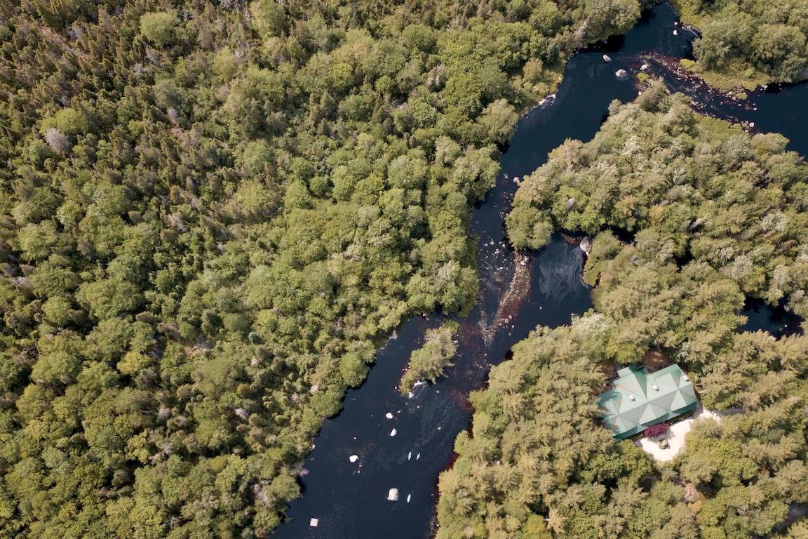 Luchtfoto van een donkere rivier die door een dicht groen bos kronkelt, met een klein gebouw aan de oever.
