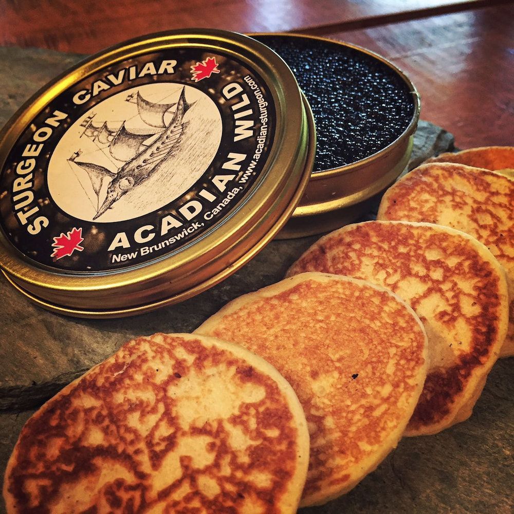 Een blikje steurkaviaar naast blini's op een houten tafel.