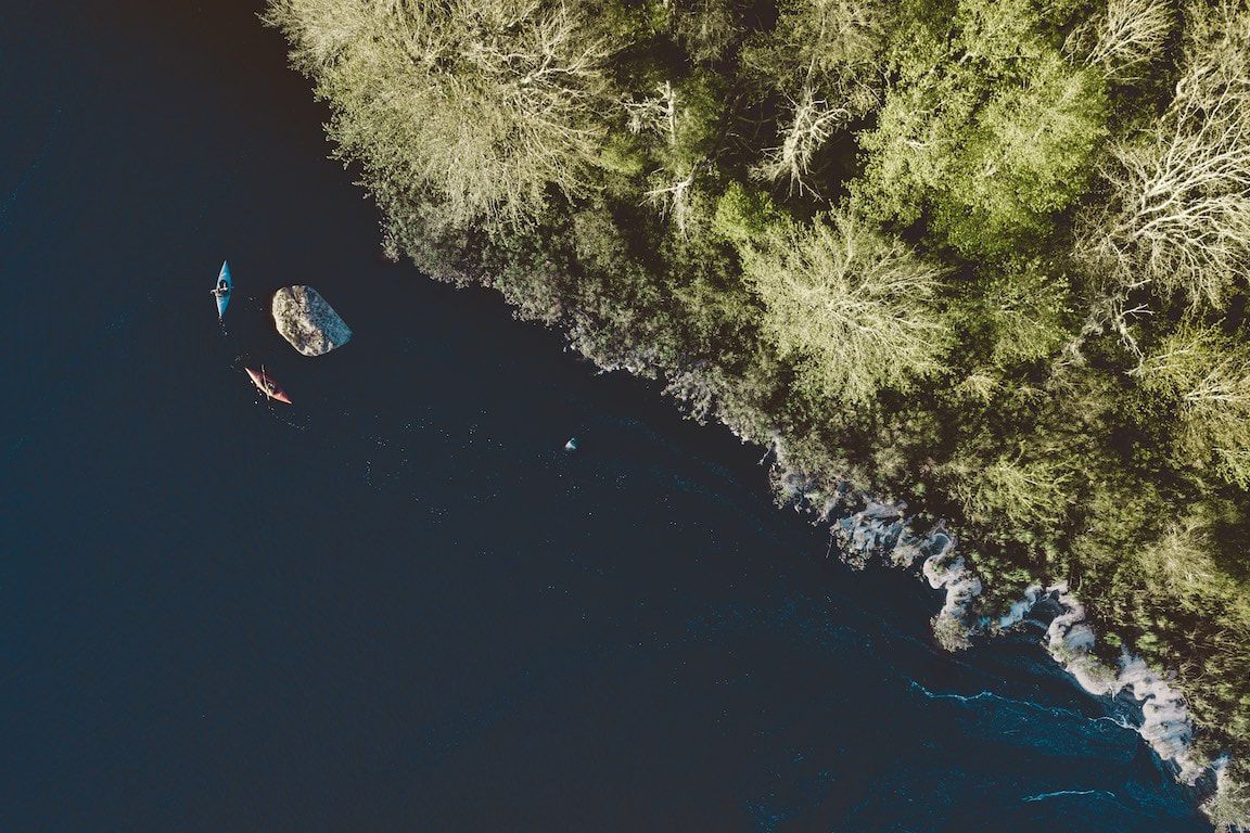 Luchtfoto van een rotsachtige kustlijn met donkerblauw water en een kleine kajak vlakbij de kant.
