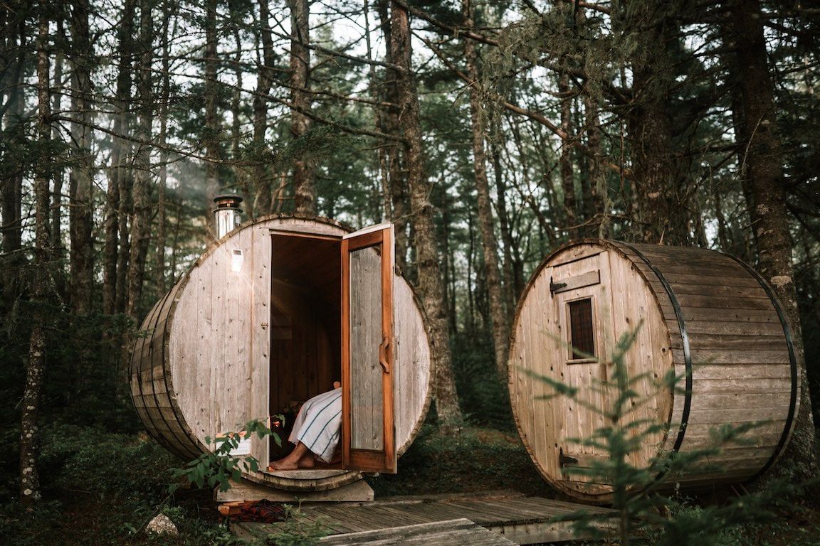 Twee houten saunatonnen in een bos, waarvan er één open is met een persoon erin.