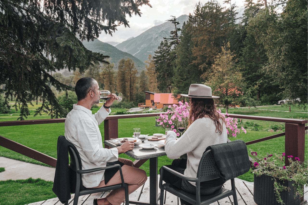 Twee mensen ontbijten op het terras van Tweedsmuir Park Lodge met uitzicht op een groene bergvallei met bomen en bloemen.