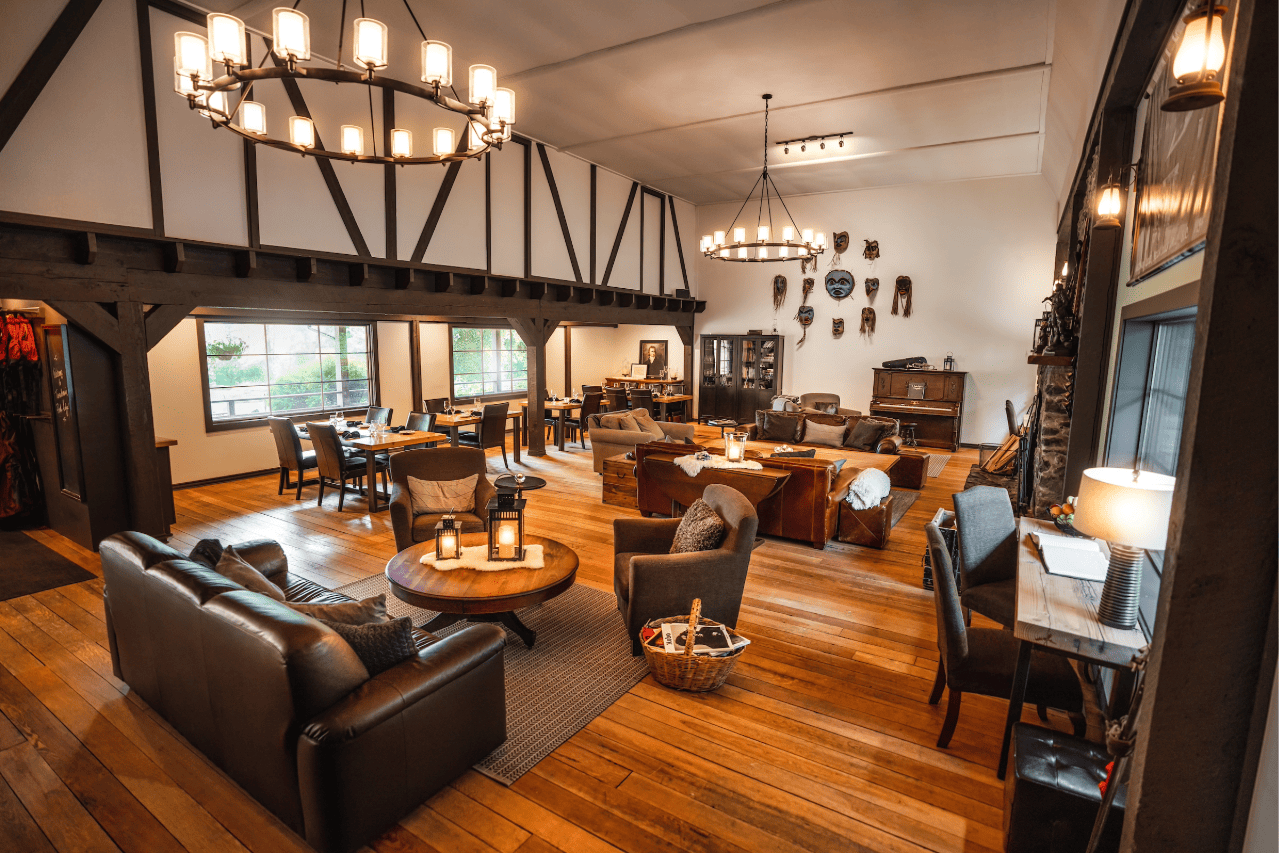 Gezellige, rustieke lounge van Tweedsmuir Park Lodge met leren zitmeubelen, houten vloeren, kroonluchters en een open haard bij grote ramen.