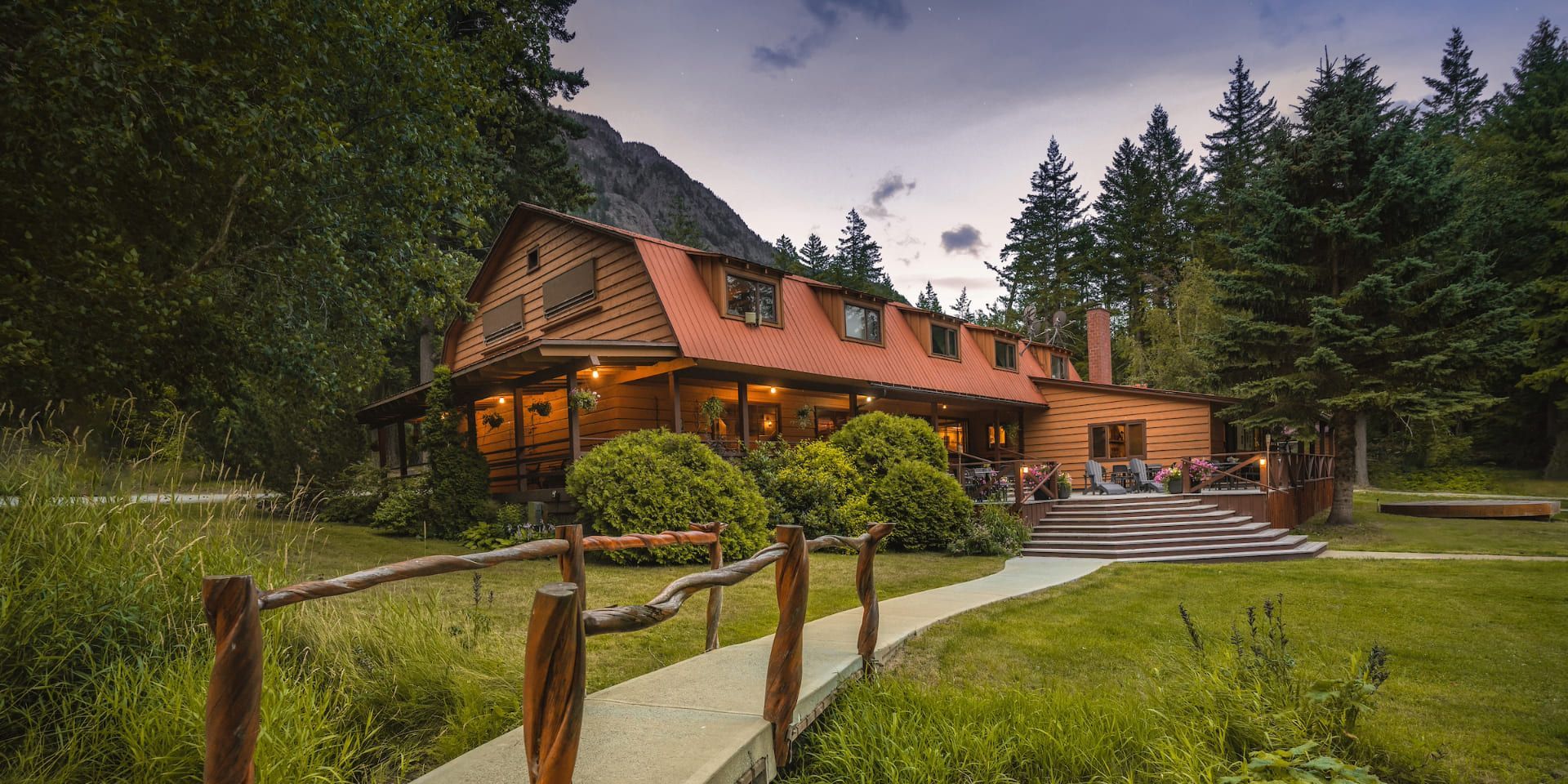 Het hoofdgebouw van Tweedsmuir Park Lodge met oranje verlichte ramen, gelegen naast een groen gazon en een bospad in de schemering.