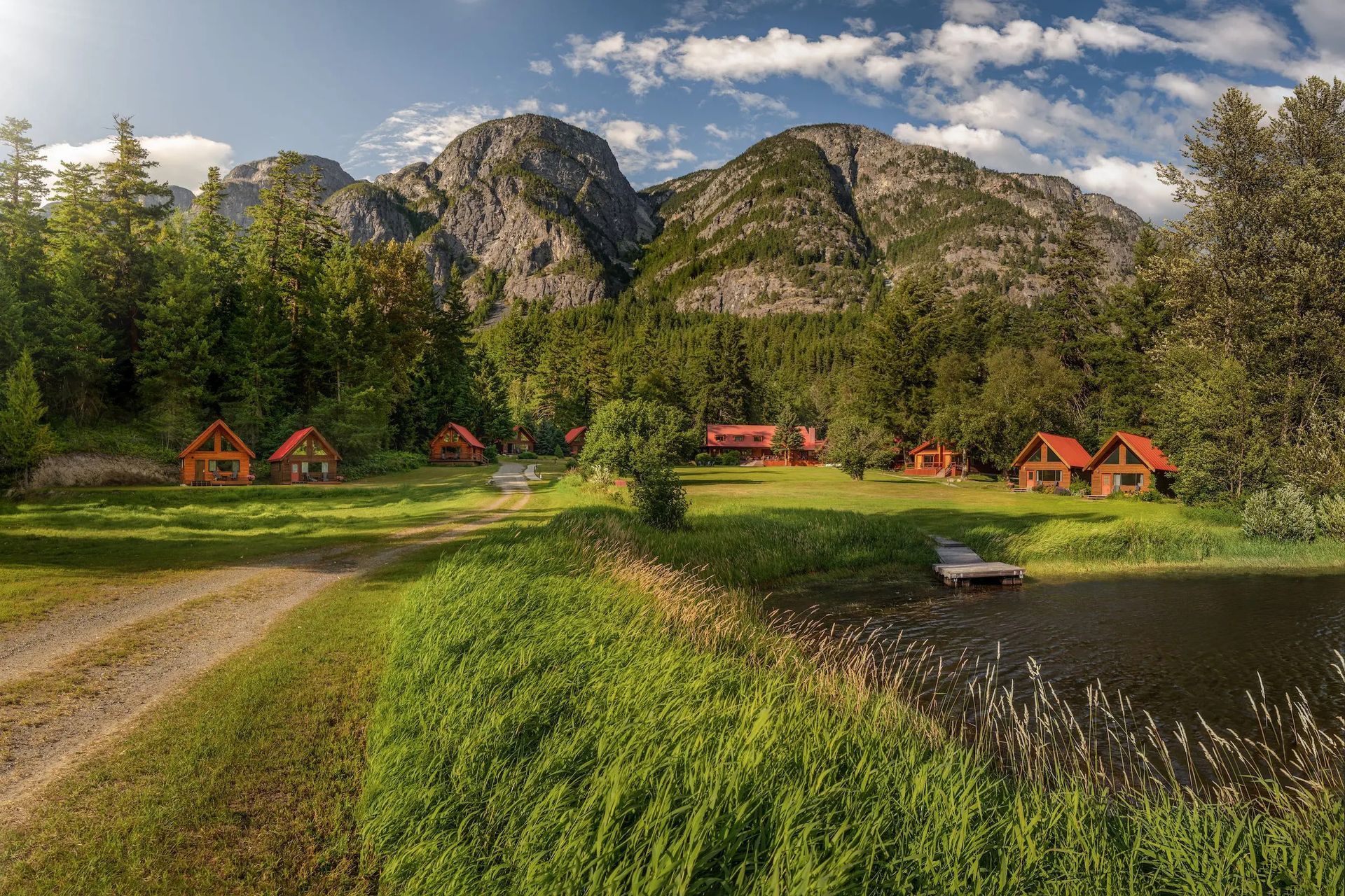 Hutten van Tweedsmuir Park Lodge aan het water, onder rotsachtige bergen, onder een gedeeltelijk bewolkte hemel.