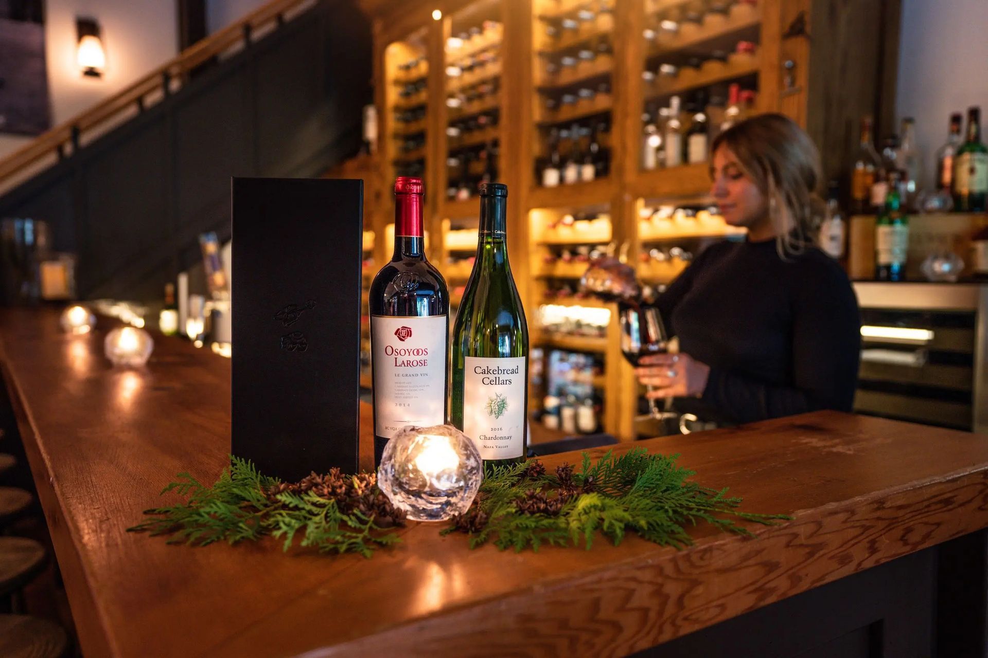 Flessen op een bar met planten en kaarslicht, barman van Tweedsmuir Park Lodge op de achtergrond.
