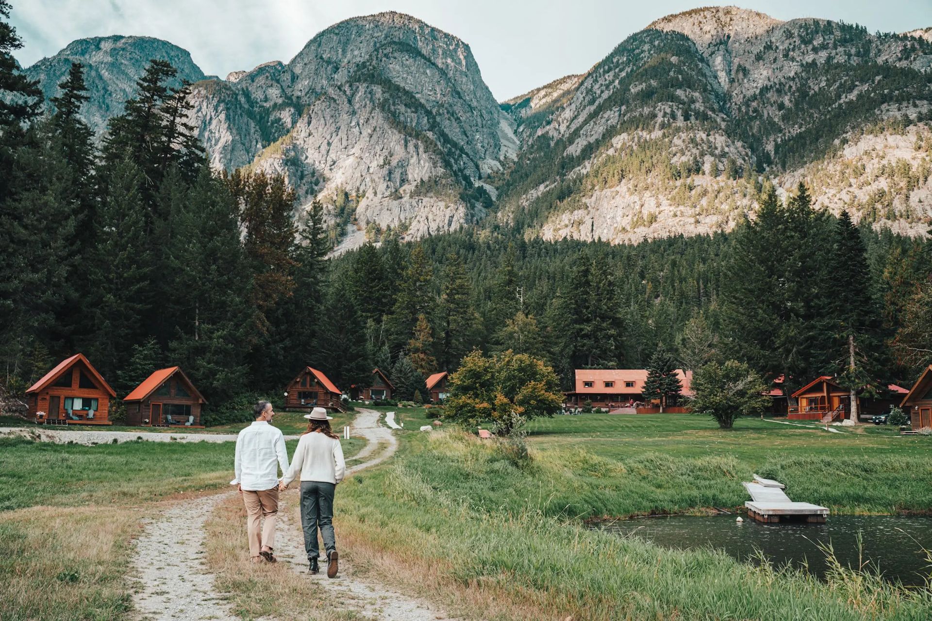Twee mensen lopen over een bospad langs hutten van Tweedsmuir Park Lodge en een meer, met bergen op de achtergrond.