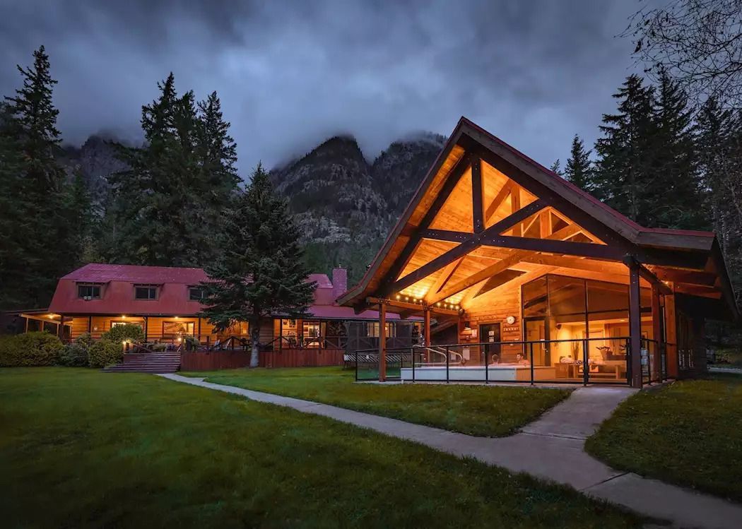 De wellness van Tweedsmuir Park Lodge die in de schemering oplicht, met dennenbomen, mistige bergtoppen en een graspad dat naar de ingang leidt.