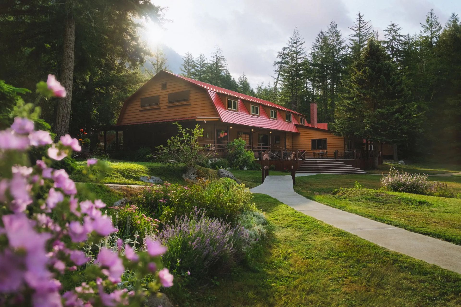 Het hoofdgebouw van Tweedsmuir Park Lodge met rood dak, verscholen tussen hoge dennenbomen, omgeven door een weelderige tuin en een kronkelend pad.