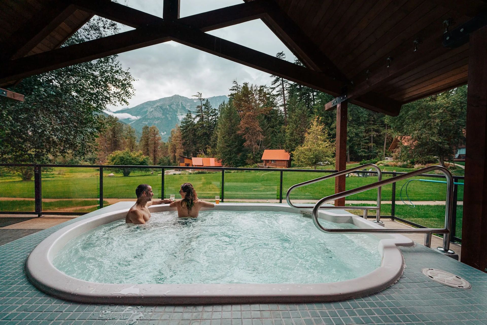 Twee mensen ontspannen in de jacuzzi van Tweedsmuir Park Lodge onder een afdak, met uitzicht op een groen berglandschap.