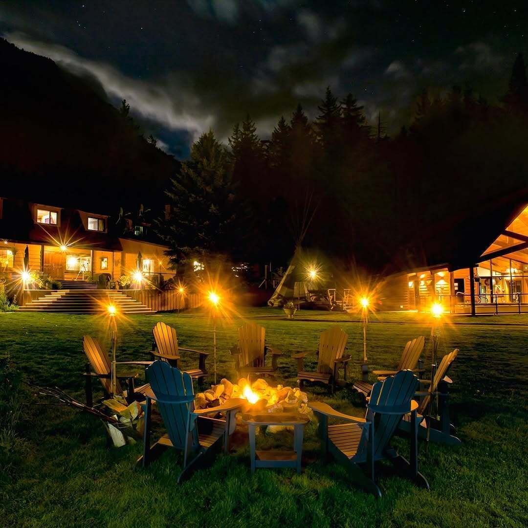 Een sfeervol vakantiehuisje van Tweedsmuir Park Lodge 's nachts met een brandende vuurplaats en Adirondack-stoelen onder een sterrenhemel.
