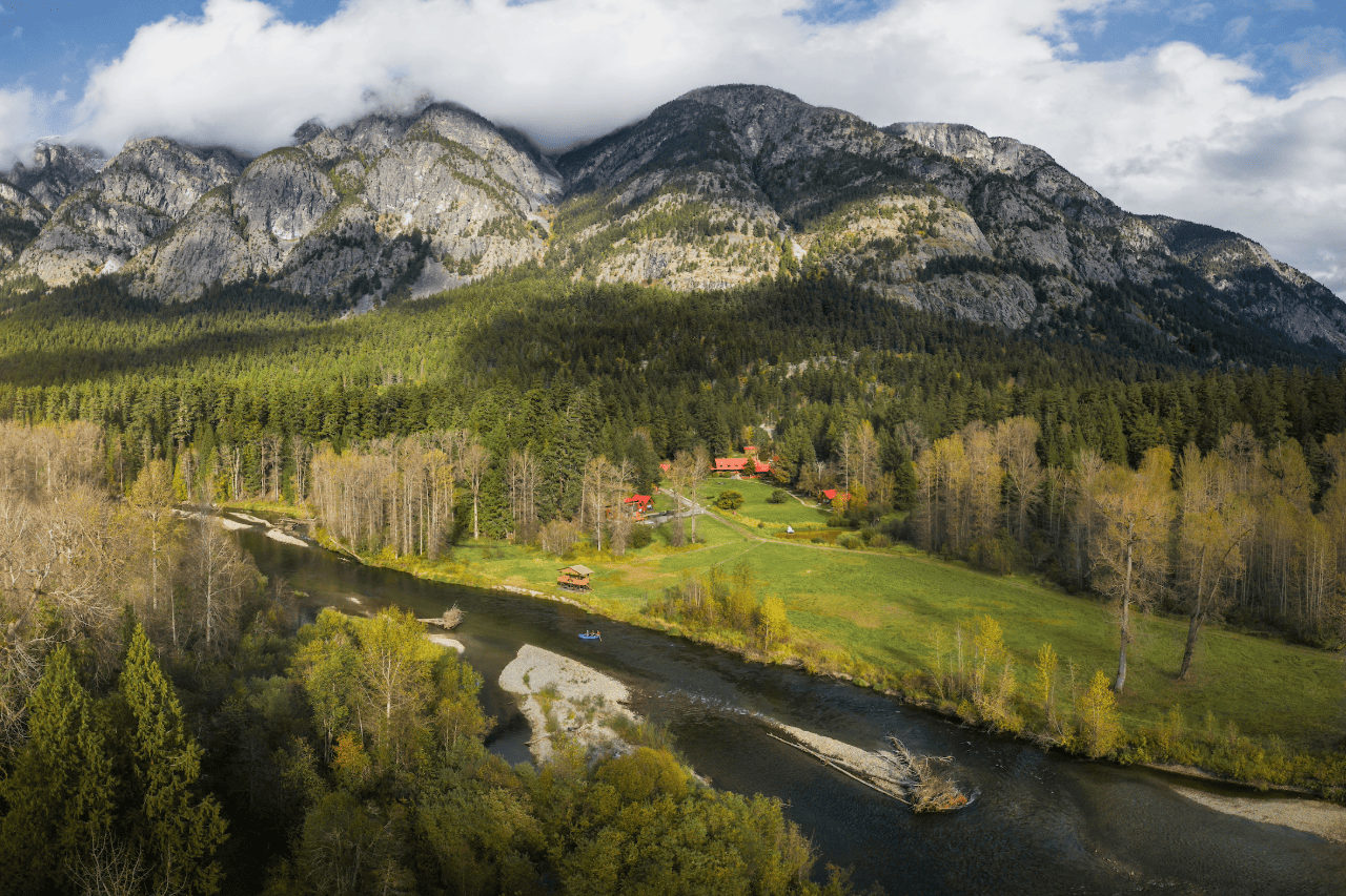 Tweedsmuir Park Lodge omgeven door een berglandschap met een kronkelende rivier, groene weide, bos en bewolkte bergtoppen op de achtergrond.