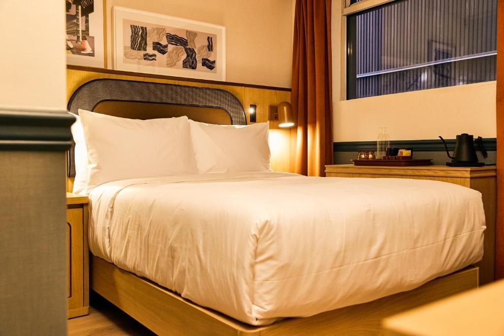 Gezellige hotelkamer met een netjes opgemaakt wit bed, een nachtlampje en een raam met jaloezieën en sfeervolle verlichting.