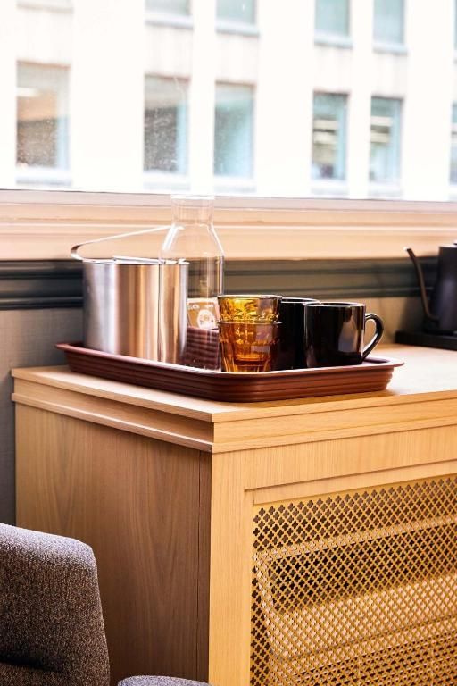 Dienblad met metalen waterkoker, amberkleurig glas en zwart kopje op een houten kast bij een raam.