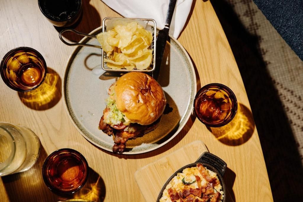 Bovenaanzicht van een hamburger, friet en drankjes op een lichte houten tafel met een bord en kleine schaaltjes.