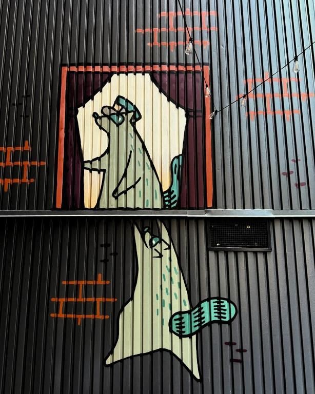 Graffiti-muurschildering van een bleke figuur op een golfplaten muur, omlijnd met zwart en oranje.