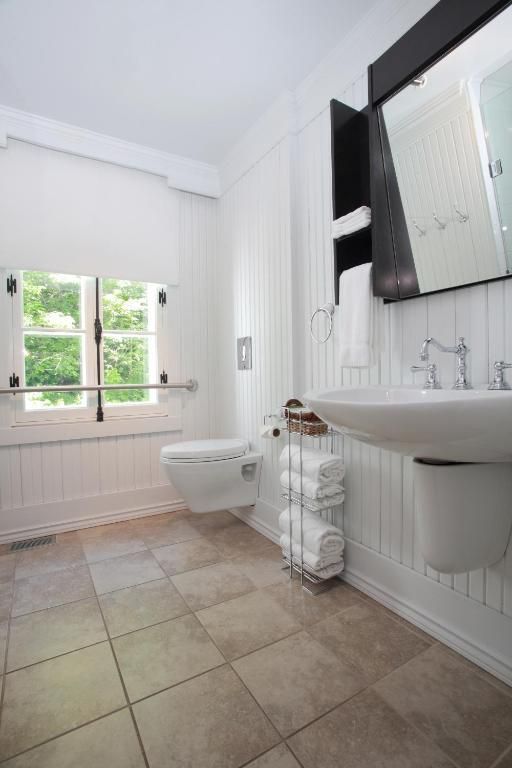 Lichte badkamer van Village Historique de Val-Jalbert met witte armaturen, tegelvloer, raam, toilet, wastafel en wandspiegel.