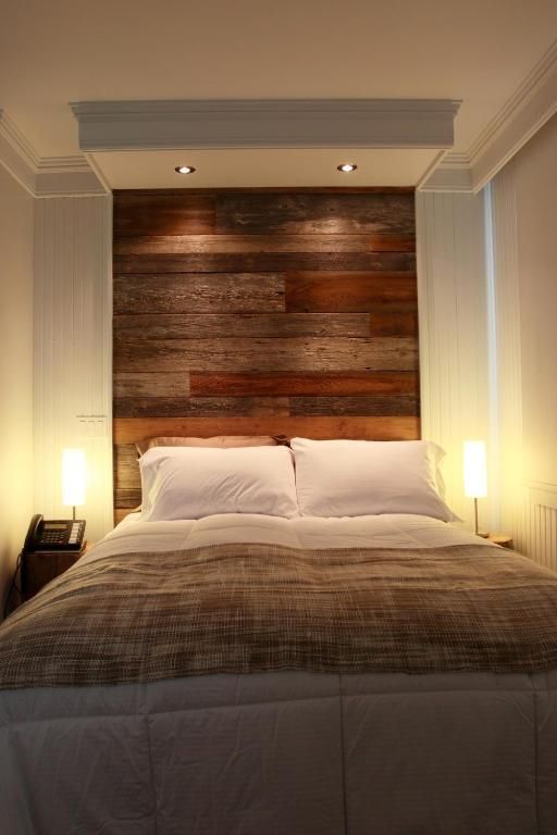 Een knus bed in het midden van een smalle slaapkamer van Village Historique de Val-Jalbert met een hoofdeinde van houten panelen en sfeervolle nachtlampjes.