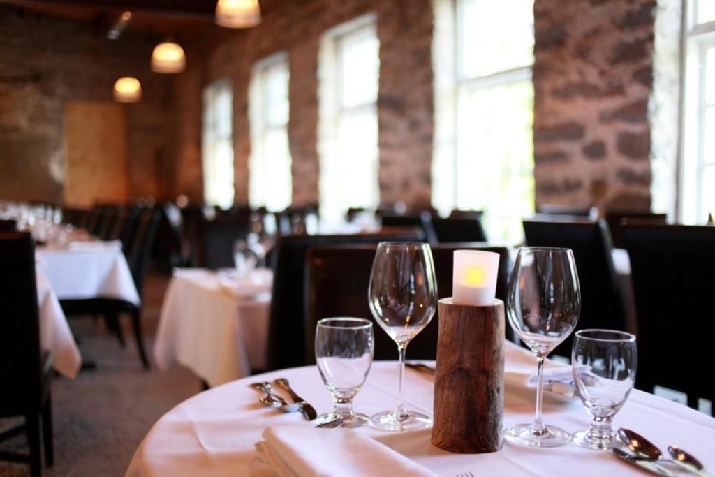 Een elegante eettafel in een restaurant van Village Historique de Val-Jalbert, gedekt met wijnglazen, kaarsen en wit tafellinnen, in een rustiek interieur.