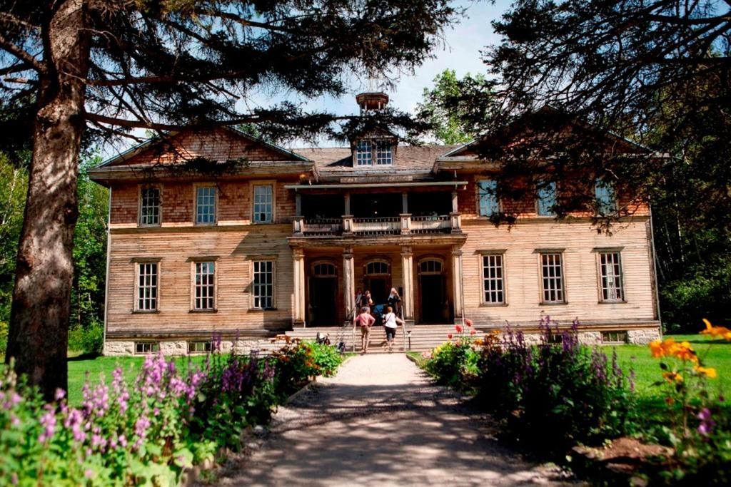 Historisch herenhuis met twee verdiepingen en een veranda, omgeven door bomen en bloemperken langs een tuinpad.