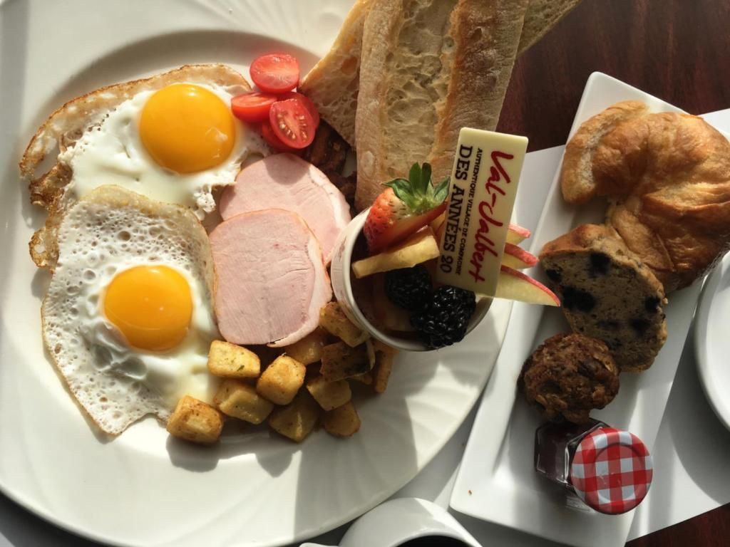 Ontbijtbord met eieren, ham, aardappelen, toast, fruit en jam, met daarnaast een croissant op een apart bordje.