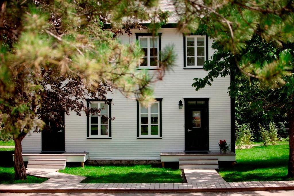 Wit, twee verdiepingen tellend huis met zwarte luiken, omgeven door bomen en een groen gazon met een oprit.