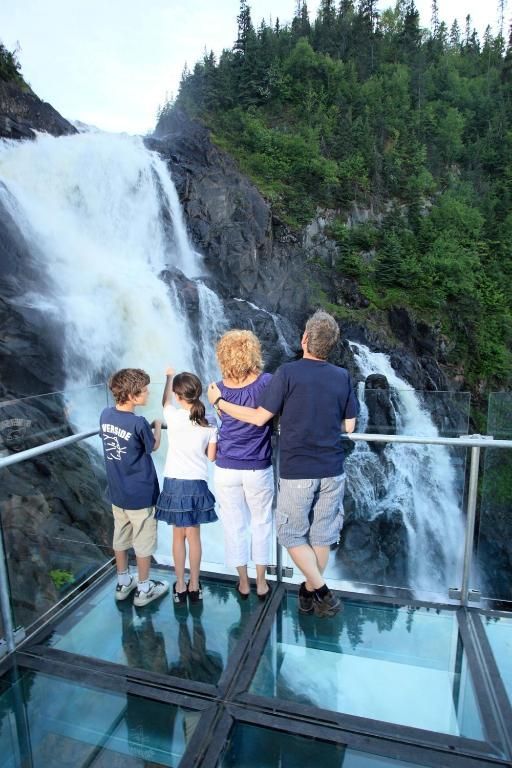 Vier mensen staan op een glazen uitkijkplatform en kijken uit op een hoge waterval in een beboste kloof.