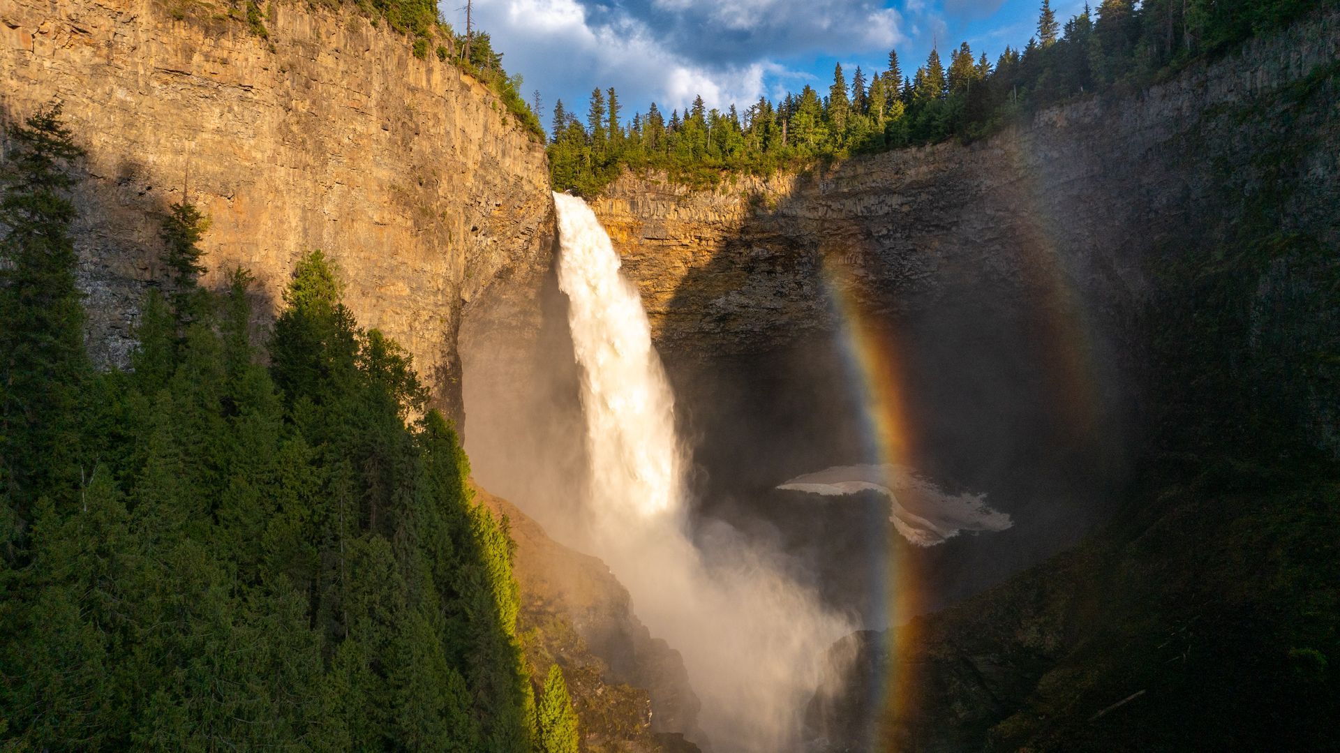 De Helckem Falls in Wells Grey Provincial Park stort neer in een beboste kloof, gehuld in mist en met een regenboog in het zonlicht.