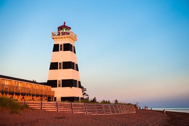 Een vuurtoren bij zonsondergang, naast een strand en een houten wandelpad onder een blauwe hemel.