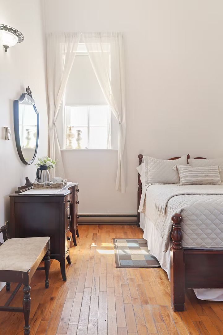 Kleine slaapkamer met een houten bed, spiegel, bankje en zonlicht dat door een raam op een houten vloer schijnt.