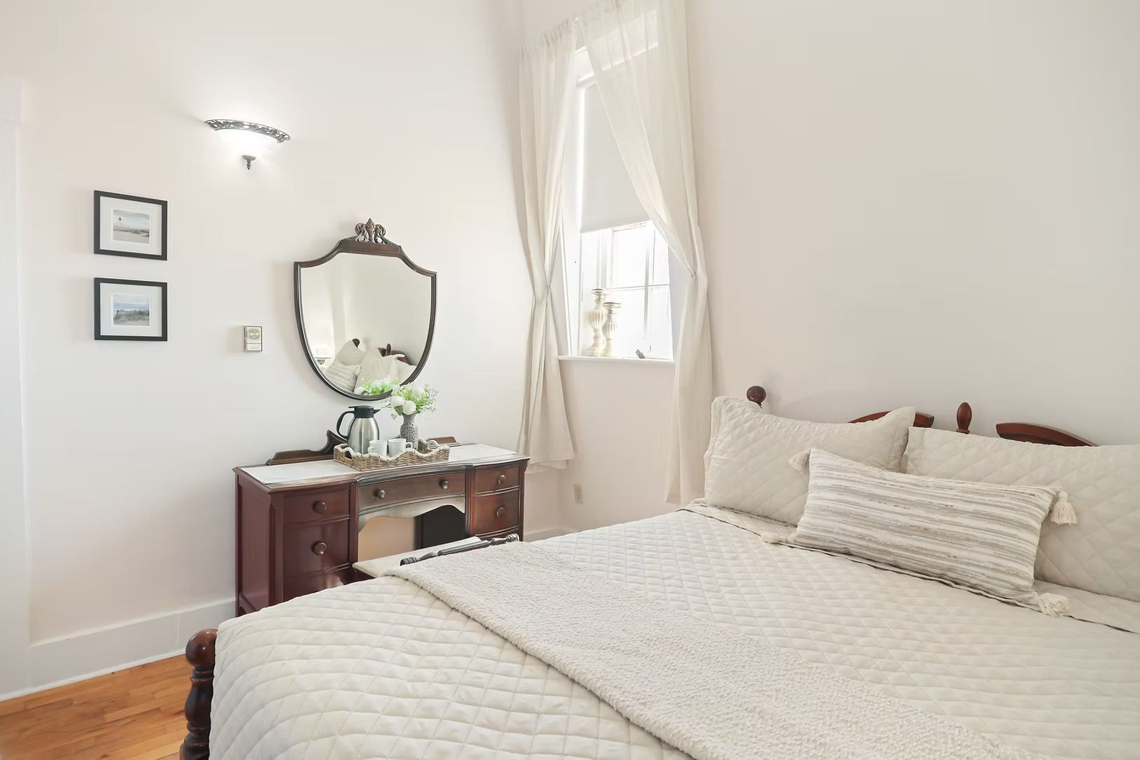 Lichte, witte slaapkamer met een bed, een vintage commode, een spiegel en een raam met transparante gordijnen.
