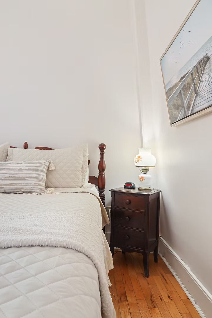 Kleine slaapkamer met een bed, nachtkastje, lamp, houten vloer en ingelijste kunst aan de muur.