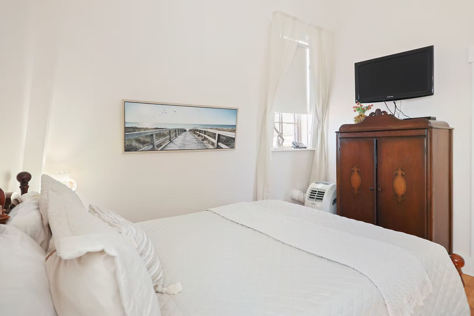 Lichte, witte slaapkamer met een bed, muurdecoratie, raam en houten kledingkast.