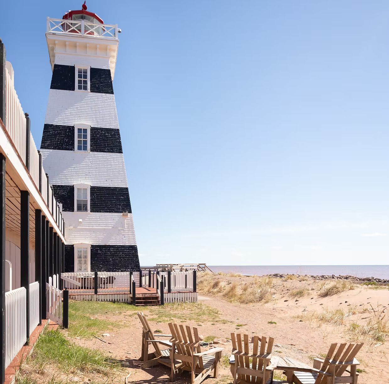 Een zwart-witte vuurtoren naast een zandstrand met Adirondack-stoelen onder een helderblauwe hemel.
