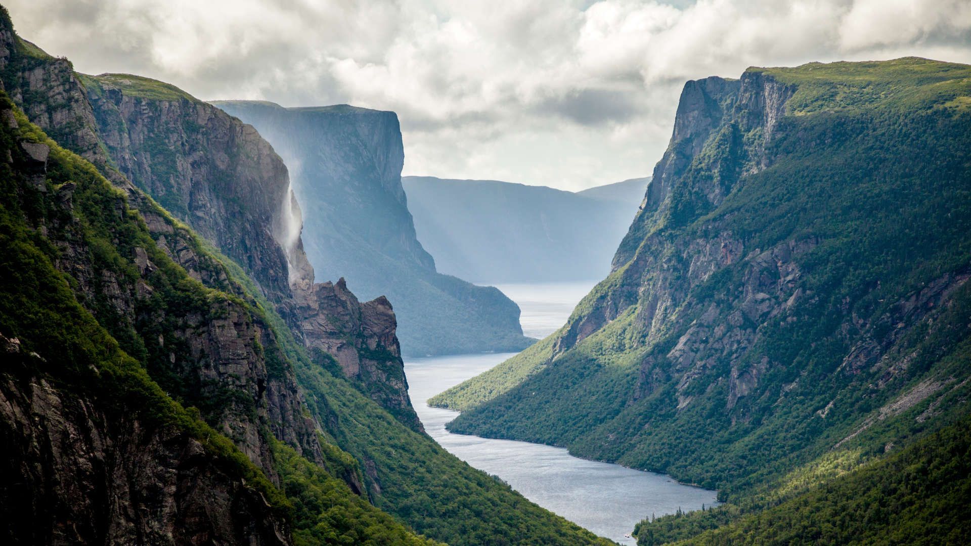 Steile, groene fjordkliffen omlijsten een kronkelende rivier onder een bewolkte hemel.