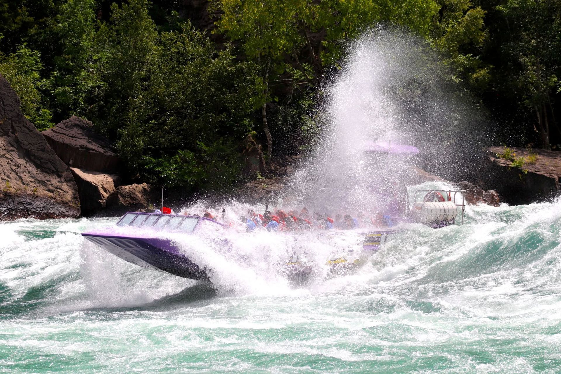 Een speedboot spuit water op terwijl hij over een rivier raast, met bomen en een rotsachtige oever op de achtergrond.