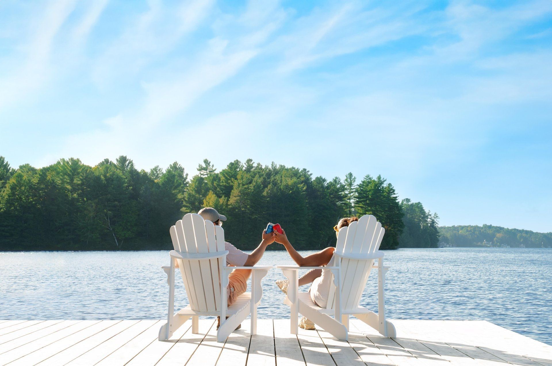 Twee mensen klinken hun glazen op witte stoelen aan een zonnig meer met een beboste oever.